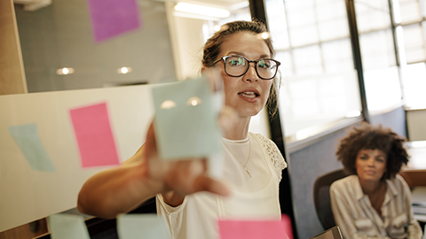 Business woman pointing at sticky note to female colleague on glass wall in office