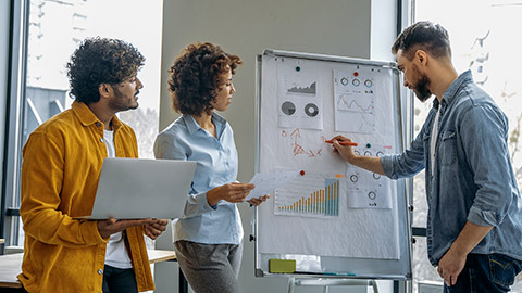 working together on a new project, brainstorming, standing near whiteboard with financial graphs, predicting income, analyzing risks, setting goals