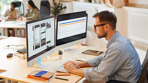 Serious programmer sitting at his workplace using computers in his work he developing new software at office with his colleagues in the background