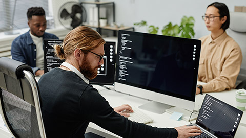 Young serious diversity programmer typing on laptop keyboard while sitting in front of computers by workplace against two coworkers