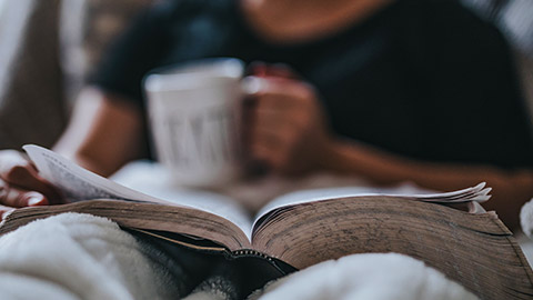 person sitting in bed reading book and drinking coffee