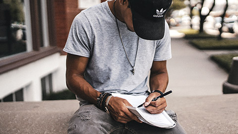 A young man sitting on wall outside a building writing in a notebook