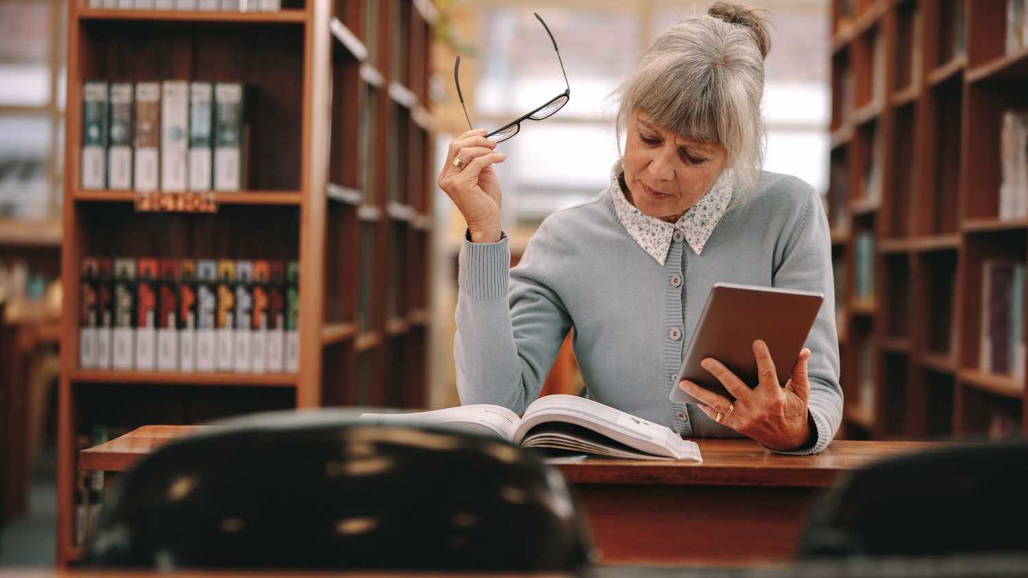 Senior woman using a tablet pc for reference while reading a book in library.