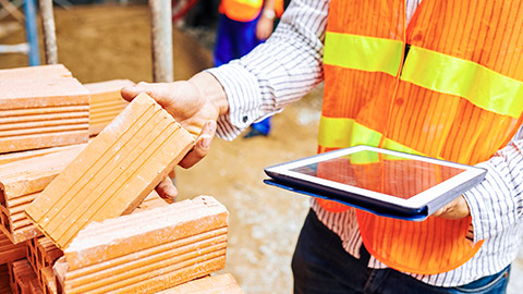 Person on building site inspecting bricks with tablet device in other hand