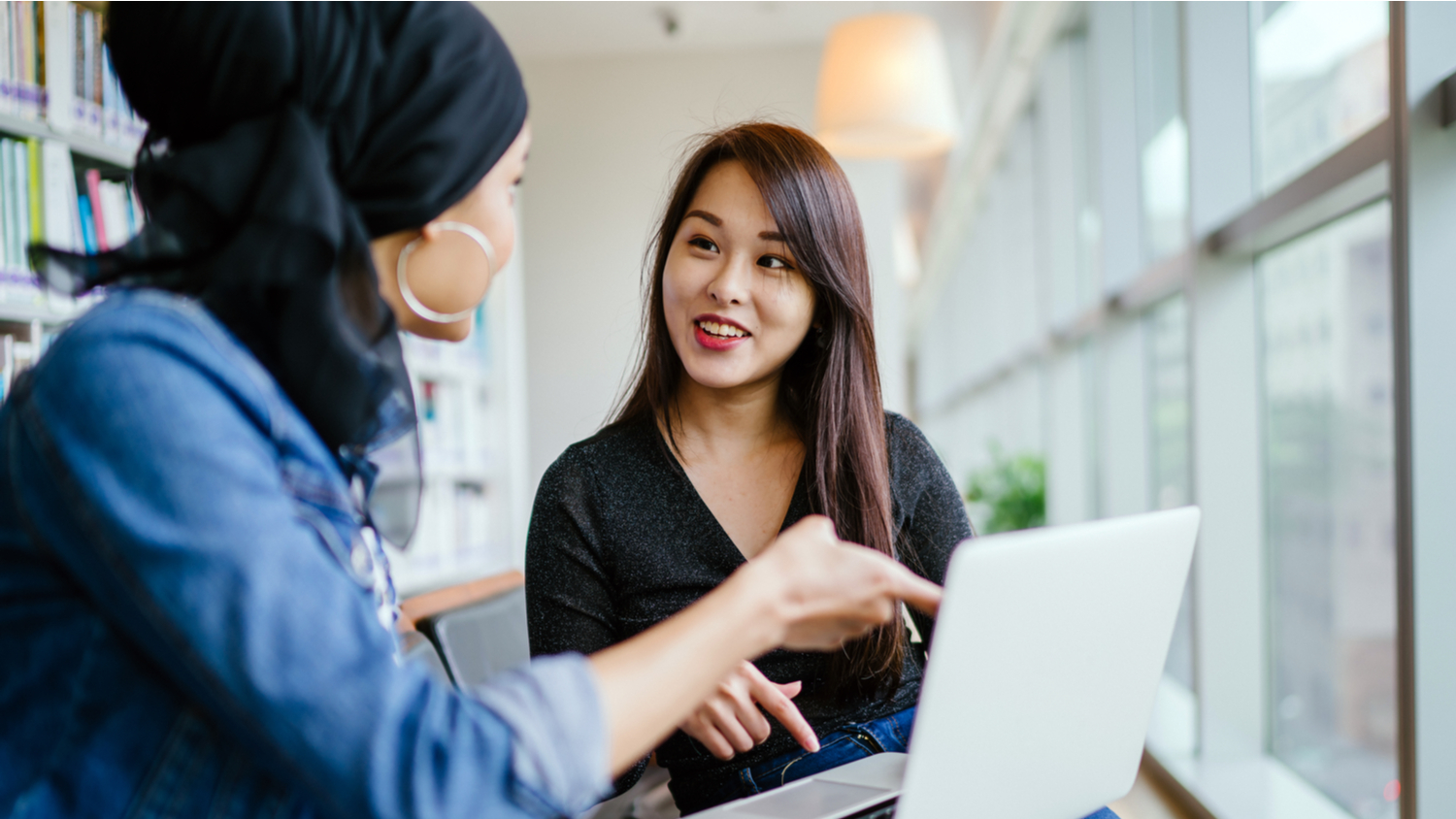 A young and Chinese Asian woman discussing feedback with a Malay Muslim woman. They are both sitting in an office and talking over a laptop.