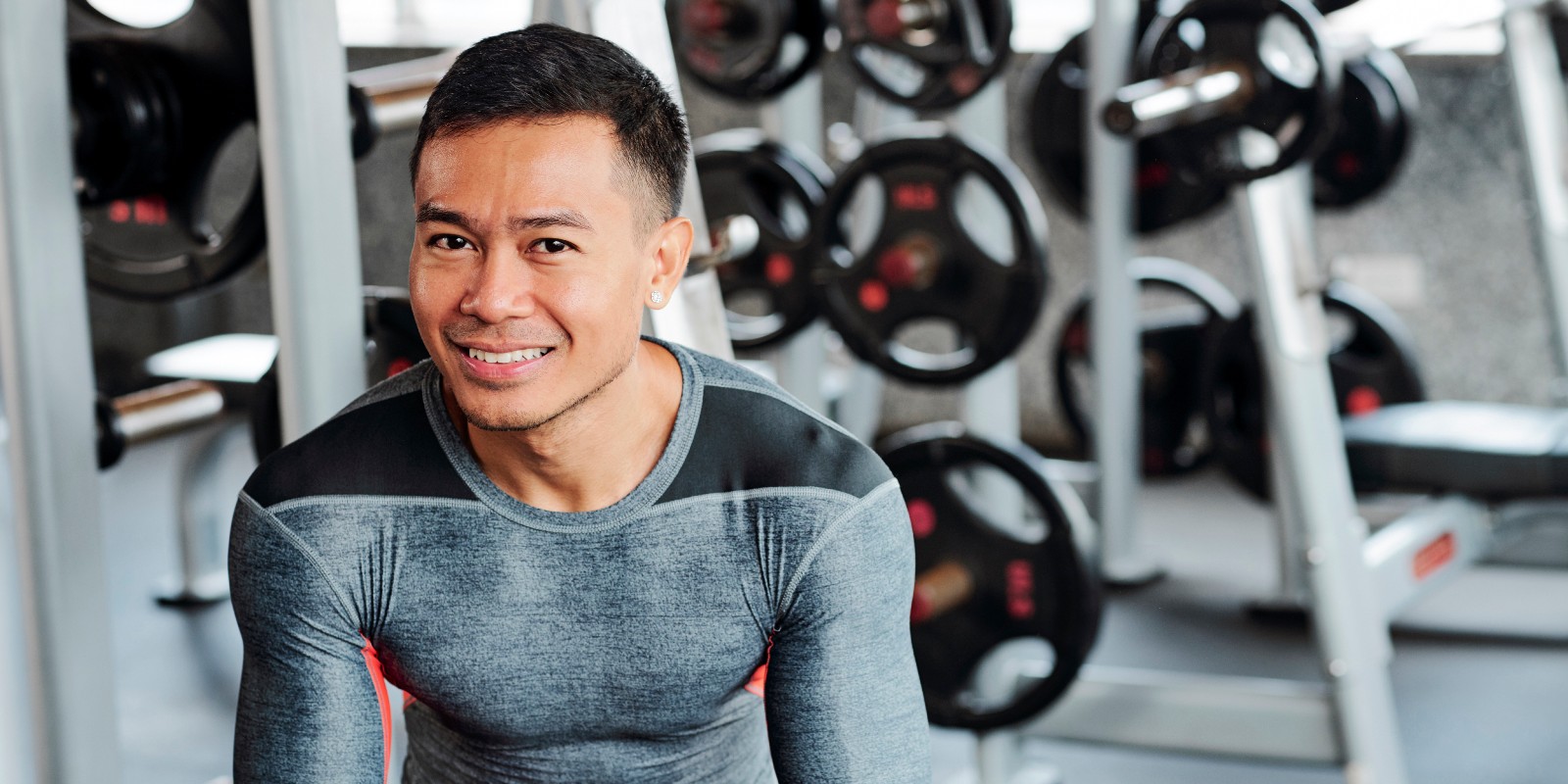 Man in fitnes attire sitting near dumbbell rack looking directly at camera and smiling