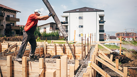 A construction worker pouring concrete into an excavation site with a neighbouring property in the background